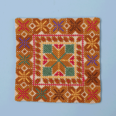 A square embroidered textile or patch coaster featuring a geometric folk-art pattern in orange, yellow, pink, green, blue, and beige on a light blue background.