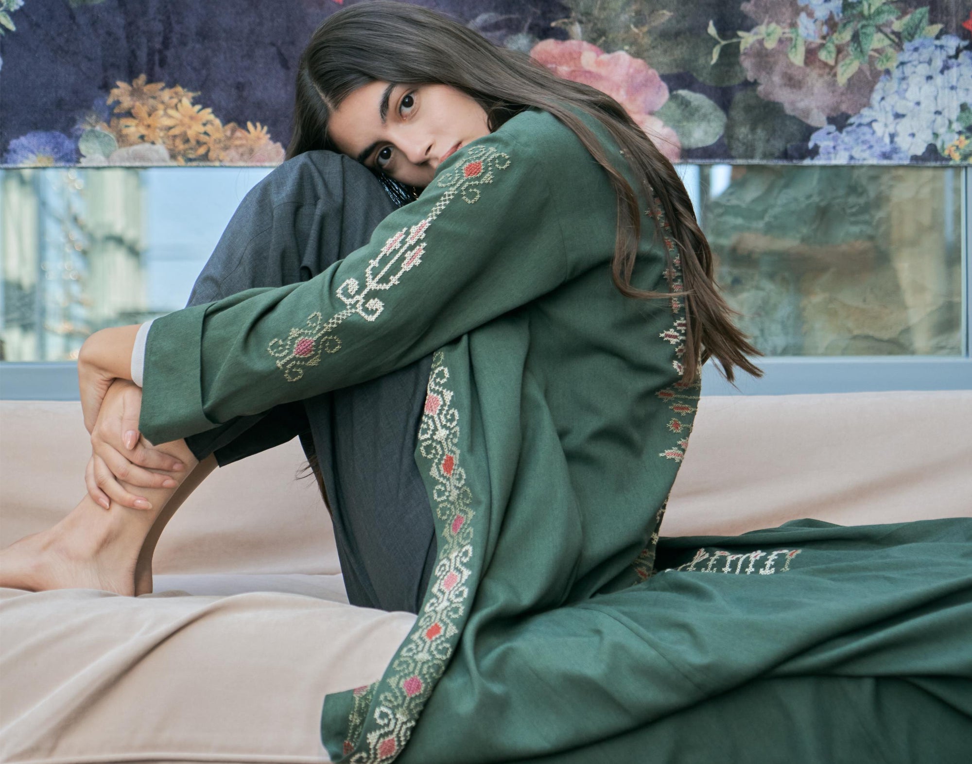 Woman in a green embroidered traditional outfit sitting on a couch with a floral wall in the background