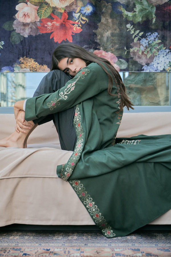 Woman in a green embroidered traditional outfit sitting on a couch with a floral wall in the background