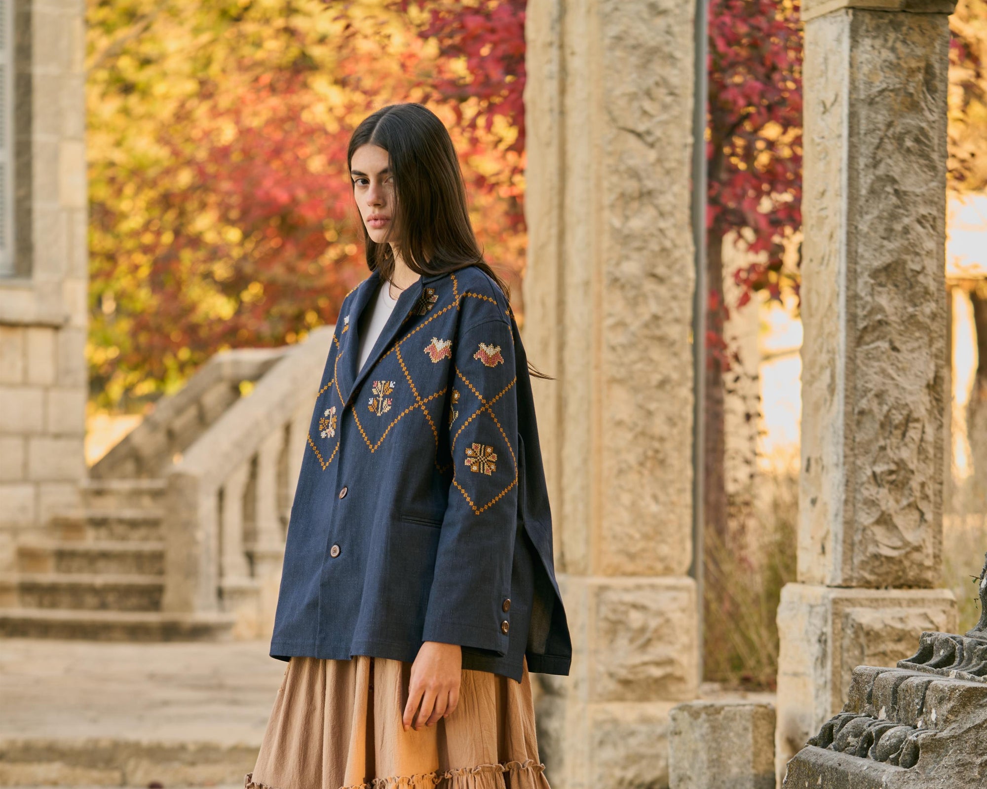Woman in a blue embroidered jacket and beige skirt standing in an outdoor setting with stone architecture and autumn foliage.