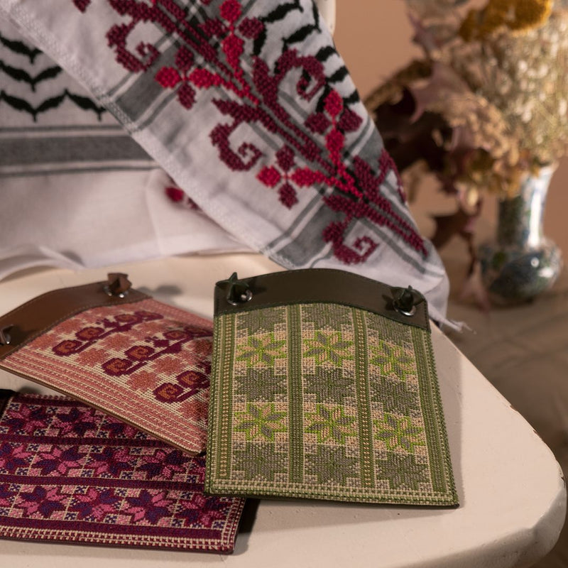 A close-up of embroidered textile samples displayed on a white chair, including a green and beige patterned fabric piece, a pink and red floral textile, and a white, gray, and burgundy embroidered cloth draped over the chair.